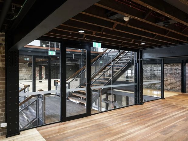 Interior view of a dramatic multi-level commercial or adaptive-reuse space, with exposed dark timber ceiling joists, black steel structural framing, extensive internal glazing, exposed brick walls, and a broad timber floor. Open metal staircases and mezzanine circulation are visible beyond the glazed partitions, creating an industrial loft-like atmosphere.