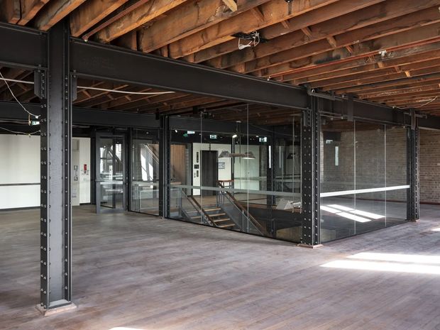 An open-plan loft-style commercial interior with exposed timber floor joists and beams, black steel structural columns and beams, exposed brick walls, and extensive internal glass partitions. A glazed stairwell and mezzanine-like circulation area occupy the centre, while the broad timber floor and industrial structural framework dominate the space.