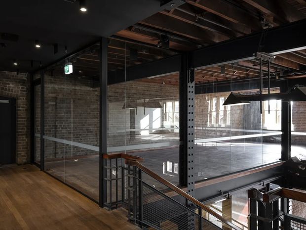 An industrial-style multi-level interior with exposed brick walls, raw timber ceiling structure, black steel columns and beams, extensive internal glass partitions, and a metal mesh stair balustrade. Warm timber flooring contrasts with the dark structural framework, while daylight enters through tall windows. The space appears to be a loft, converted warehouse, or commercial workspace with an open stair and mezzanine area.
