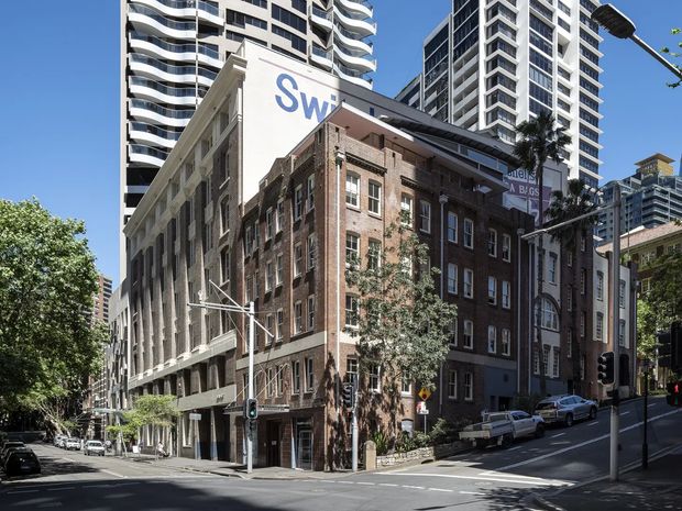 Street-level view of a dense urban intersection featuring a retained historic brick masonry building with arched and rectangular windows, integrated with a contemporary multi-storey addition and surrounded by tall modern apartment or commercial towers. Mature street trees, traffic signals, sloping roads and urban streetscape are visible under clear daylight.