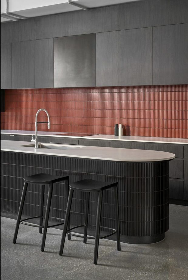 A sophisticated contemporary kitchen featuring a large rounded island with a pale stone benchtop and vertically fluted dark cabinetry. Two black bar stools sit at the island. The rear wall combines dark wood-look cabinetry, a stainless-steel rangehood, and a striking red-brown vertically ribbed tile splashback. A curved chrome tap and integrated sink complete the refined, high-contrast design.
