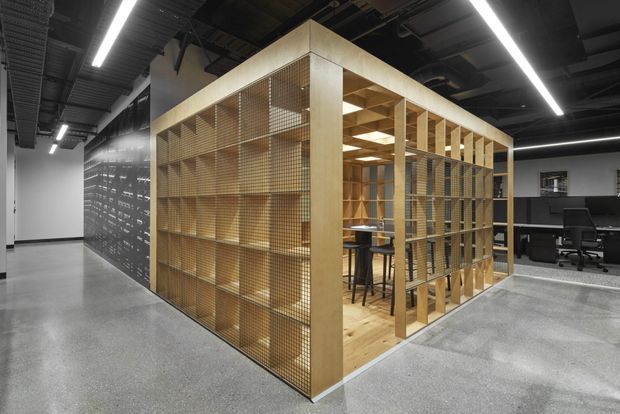Contemporary commercial office interior featuring a freestanding timber-framed enclosure with fine metal mesh shelving or screening. The structure forms a semi-enclosed meeting or breakout area with high stools and a table, while open-plan workstations are visible behind it. The space has polished concrete flooring, exposed dark ceiling services, linear LED lighting, and a monochrome industrial palette accented by warm timber.