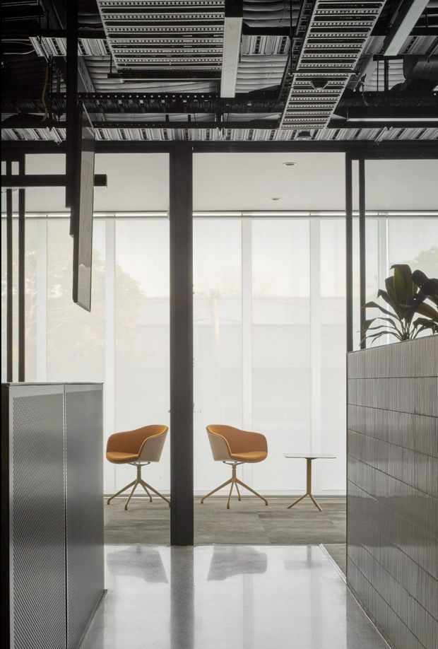 A refined commercial office interior with exposed black structural framing and open services above, full-height translucent roller blinds, polished concrete flooring, and a small seating area with two tan lounge chairs and a side table. Perforated metal cabinetry and a tiled partition with integrated planting frame the space, creating a contemporary industrial workplace.
