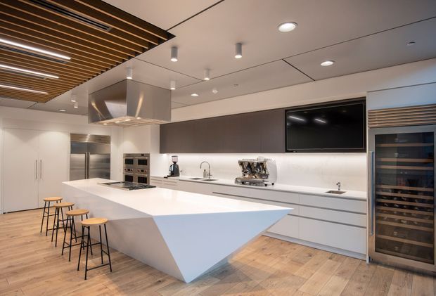 Contemporary open-plan kitchen with a large sculptural white island, integrated appliances, extensive white cabinetry, dark wall-mounted television, stainless-steel rangehood, and a built-in wine refrigerator. Warm timber flooring contrasts with the crisp white and charcoal finishes. A slatted timber ceiling feature with integrated linear lighting adds visual interest.