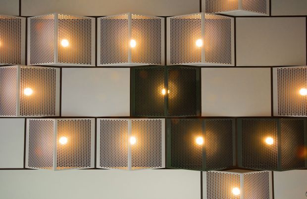A close-up of a geometric interior wall installation composed of rectangular perforated metal panels arranged in a staggered grid. Warm glowing lights shine through the perforations, creating a patterned, atmospheric feature surface with neutral grey, cream, and dark tones.