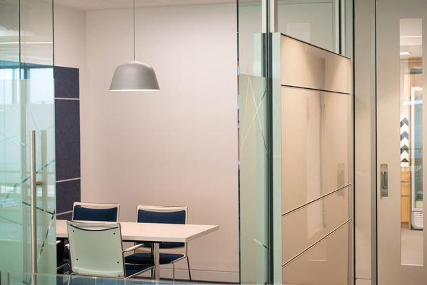 A compact contemporary office meeting or dining area enclosed by extensive clear glass partitions. A white rectangular table with modern chairs sits beneath a grey pendant light, with pale walls, dark blue accent panels, and a large glossy light-coloured built-in partition or storage wall. Additional office spaces are visible through the glazing.