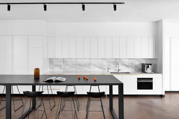 Bright, minimalist open-plan kitchen and dining area with extensive white flat-panel cabinetry, a grey marble-look backsplash, dark island bench, black bar stools, track lighting, integrated appliances, and warm herringbone timber flooring.