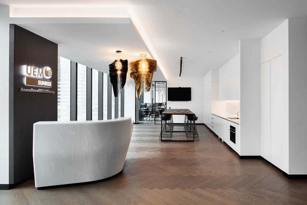A refined open-plan interior combining a reception or entry area with a contemporary kitchen and dining space. The room features a curved white reception desk, dark timber feature wall with illuminated signage, sculptural pendant lights, full-height glazing, white handleless cabinetry, integrated appliances, and a warm herringbone timber floor.