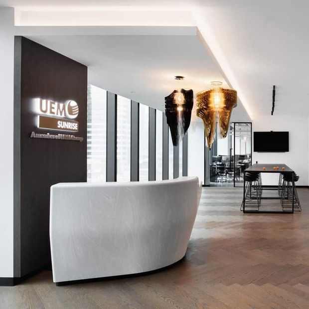 Contemporary commercial reception area with a curved white reception desk, dark timber feature wall and illuminated signage, floor-to-ceiling windows, sculptural pendant lights, timber parquet flooring, and a casual meeting area with a wall-mounted screen and table.