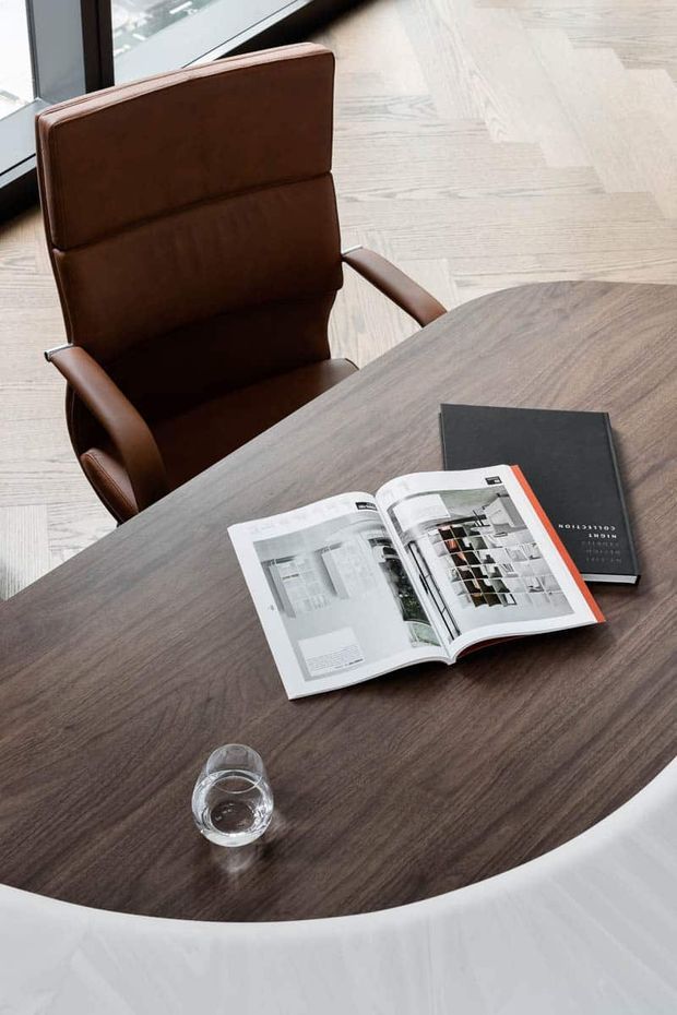 Close-up of a refined office or meeting area featuring a round dark wood meeting table, a brown leather upholstered armchair, a glass of water, and open design magazines. Light timber herringbone flooring and black-framed glazing create a contemporary professional setting.