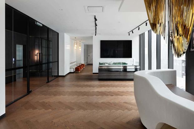 A sophisticated open-plan interior featuring a reception or lounge area with warm herringbone timber flooring, dramatic black-framed glazed cabinetry or partitions, a black media wall, and a curved white reception desk or sofa in the foreground. Large windows provide abundant daylight, while a sculptural gold pendant installation adds a luxurious focal point. Track lighting and restrained black-and-white finishes create a refined contemporary atmosphere.