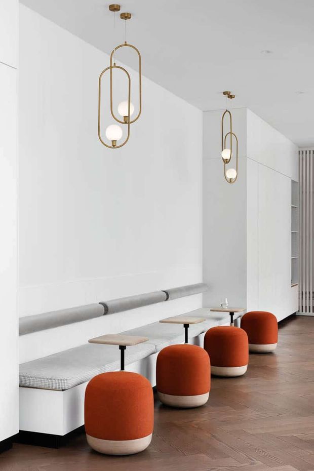 A refined minimalist seating area with a long built-in upholstered bench, small individual tables, and rounded orange-and-cream stools. The bright white interior features full-height built-in cabinetry, sculptural brass pendant lights, and warm herringbone timber flooring.