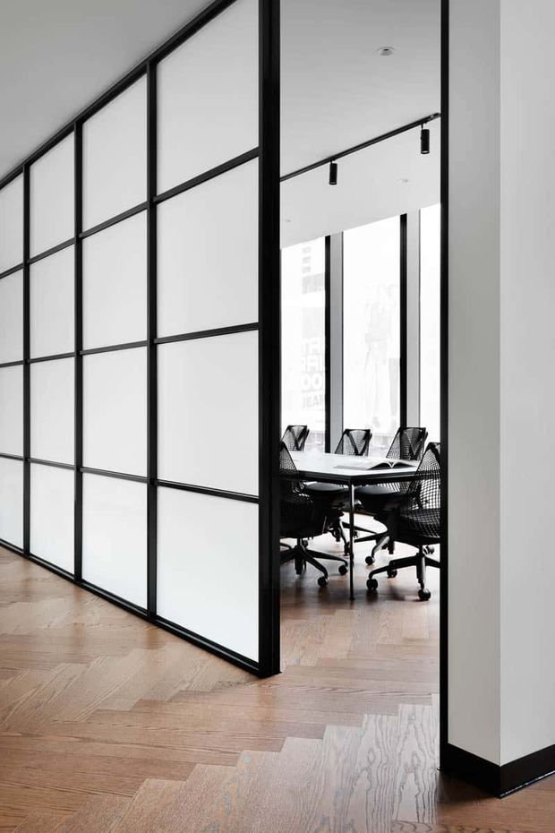 A contemporary office interior viewed through a black-framed glazed partition. The foreground features a large grid of translucent or frosted panels, while the adjoining meeting room contains a white conference table and black mesh office chairs. Pale timber parquet flooring, white walls, black skirting, and minimalist black track lighting create a clean, refined workspace.