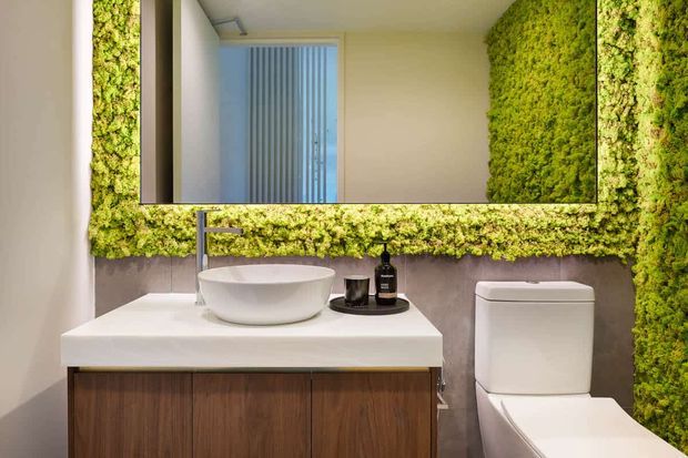 Contemporary bathroom vanity with a white vessel basin, wall-mounted chrome tapware, timber cabinetry, and a white toilet. A large mirror is framed by a lush preserved moss or vertical green wall feature, creating a strong natural accent against muted wall finishes.