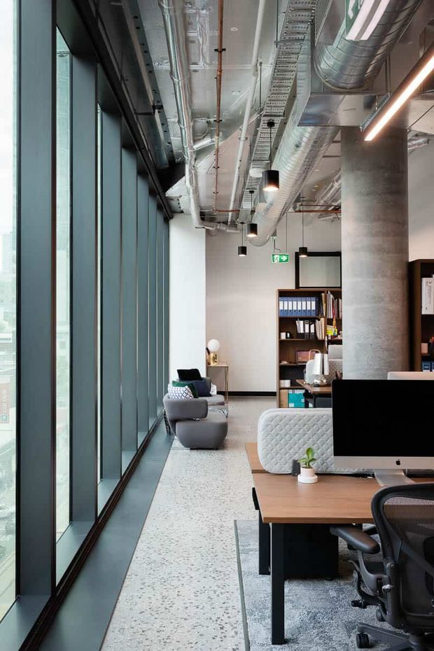 A contemporary open-plan office interior with floor-to-ceiling glazing along one side, exposed services and ductwork, concrete columns, polished terrazzo-style flooring, and modern workstations. Shelving, filing binders, computers, task chairs, and lounge seating create a functional yet refined workplace environment.