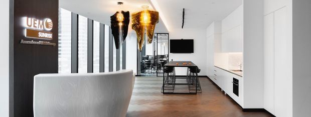 A bright contemporary commercial interior, likely a showroom or office reception and kitchen area. The space features a curved white reception counter in the foreground, dark timber wall signage, tall black-framed windows, polished timber flooring, sculptural metallic pendant lights, and a long white built-in kitchenette with integrated appliances. A meeting or dining table is visible toward the rear.