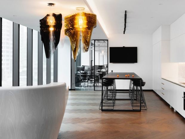 A contemporary open-plan kitchen and dining area with a compact black island table, bar stools, white built-in cabinetry, and warm herringbone timber flooring. Dramatic sculptural pendant lights hang above the foreground seating area, while full-height glazing and a glazed partition reveal an adjoining workspace or dining area. The palette is predominantly white, black, warm wood, and metallic gold.