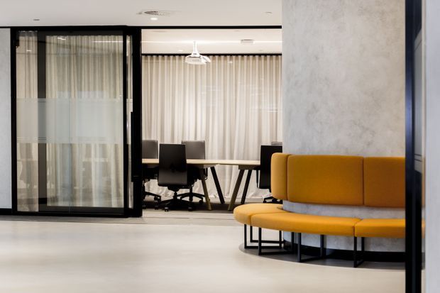 Contemporary commercial office interior with a bright open-plan meeting or collaboration area. A mustard-yellow upholstered bench wraps around a grey base in the foreground, while a meeting table and black office chairs sit behind glazed black-framed partitions with sheer curtains. The space has pale flooring, a white ceiling, and a light grey textured feature wall.