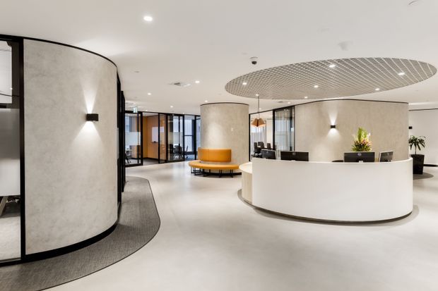 A bright contemporary commercial reception lobby with a curved white reception desk, rounded textured plaster walls, black-framed glazed partitions, an orange circular bench, and a distinctive oval suspended grid ceiling feature. The space has polished light flooring, integrated wall lighting, and a clean, refined office or hospitality feel.