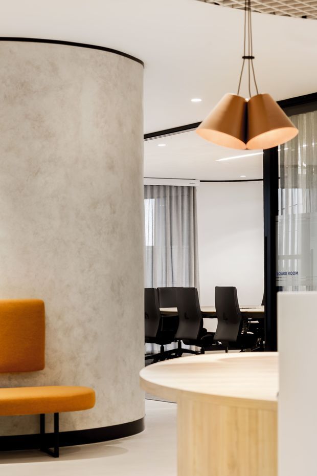 Contemporary commercial office interior featuring a curved textured concrete partition, built-in mustard upholstered seating, a rounded light timber reception or meeting table, black-framed glazed meeting room, and a sculptural warm-metal pendant light. A boardroom with dark office chairs is visible beyond the partition. The space has a refined neutral palette with black detailing and warm amber accents.
