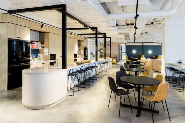 A bright contemporary commercial hospitality or workplace breakout space with an open kitchen and café-style dining area. The room features a long pale timber-clad kitchen wall, a curved white service island, extensive bar seating, round dining tables, black metal-framed furniture, exposed white ductwork, polished concrete flooring, and black-framed glazed partitions. The palette is light timber, white, black, and grey with warm yellow lighting accents.