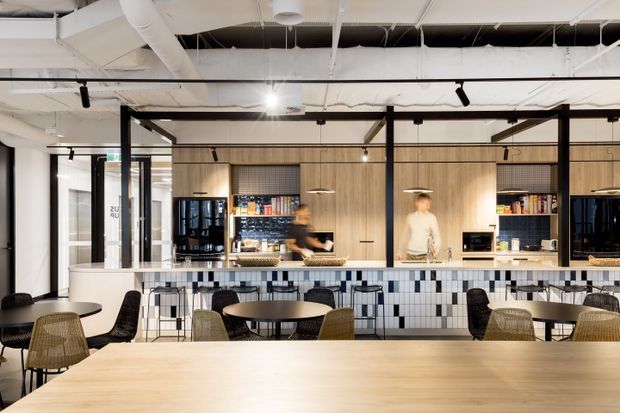 Contemporary commercial hospitality interior featuring a large communal dining table in the foreground, café-style tables and chairs, and a long service counter with a patterned tile front. Behind the counter are warm timber storage and kitchen cabinetry, shelving, appliances, pendant lighting, and exposed ceiling services with black track lighting. The open, casual fit-out has a neutral palette with timber, white, black, and blue-grey accents.