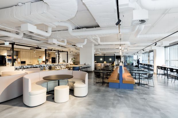 A spacious contemporary commercial interior with an open-plan café or workplace hospitality area. The room features polished concrete flooring, exposed white services and ductwork, black track lighting, extensive glazing, a curved cream upholstered booth with matching stools, communal timber-and-blue seating, and a large open kitchen or service counter at the rear.