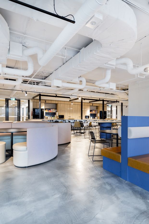 Bright open-plan commercial interior with exposed white mechanical services and ductwork, polished grey concrete flooring, black-framed glazing and partitions, timber-toned wall cabinetry, a kitchenette or refreshment counter, and varied lounge and dining seating. The space combines curved upholstered booths with vivid blue and mustard seating accents in a contemporary workplace or hospitality setting.