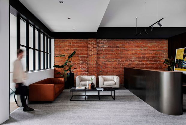 A contemporary commercial reception lounge with exposed red brick walls, black-framed floor-to-ceiling windows, a dark curved reception desk, leather and upholstered lounge seating, a low black metal coffee table, and track lighting. The space has a refined industrial character with a grey carpet and a monochrome, warm-toned palette.