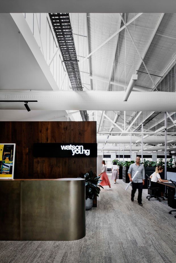 A bright contemporary commercial office reception and open-plan workspace. The image features a curved brushed-metal reception desk in front of a dark timber feature wall with a visible “watson young” sign. Exposed white-painted roof trusses, services, ducts, cable trays and high industrial ceilings dominate the upper space, while workstations, office staff and indoor plants occupy the background. Grey carpet covers the floor.