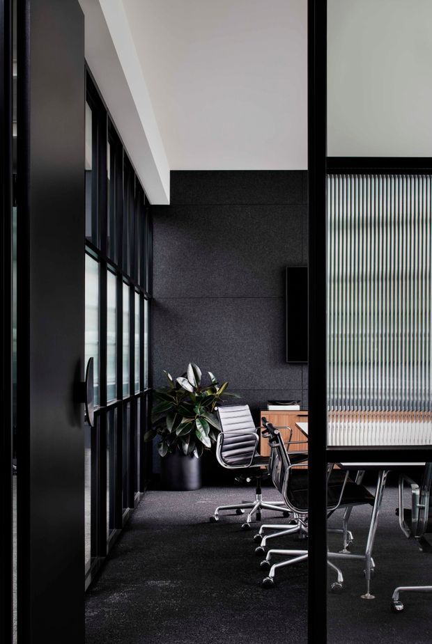 A dark, refined modern office meeting room viewed through black-framed glazed partitions. The space features charcoal textured wall panels, dark carpet, a timber meeting table, ergonomic rolling office chairs, tall black-framed windows, and a vertical ribbed translucent glass screen. A potted plant provides the main natural accent, while a white ceiling plane adds contrast.