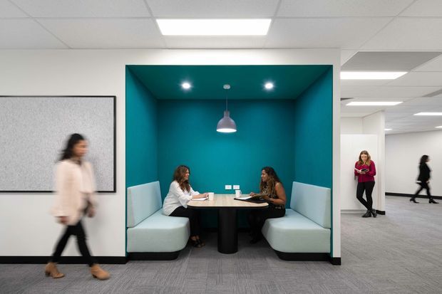 Contemporary commercial office interior featuring a recessed teal meeting booth with built-in upholstered bench seating, a round table, pendant light and downlights. The booth opens onto an open-plan office with carpet flooring, suspended acoustic ceiling tiles, fluorescent lighting and white walls. People are present in the office, including blurred passers-by.