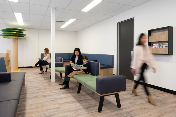 A bright, contemporary commercial waiting area with modular upholstered seating in navy and muted green, pale wood-look flooring, white walls, and a suspended grid ceiling with linear fluorescent lighting. Several people occupy or pass through the space, while a wall-mounted display shelf provides a small decorative and informational feature.