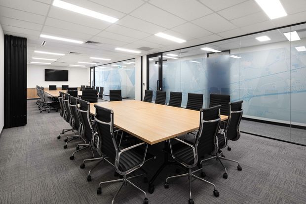 A bright contemporary commercial boardroom with a large rectangular timber meeting table surrounded by black rolling office chairs. The room has grey carpet flooring, white acoustic ceiling tiles with recessed linear lighting, glazed partitions with frosted graphic panels, and a wall-mounted display screen. Black curtains and glass-framed meeting spaces are visible in the background.
