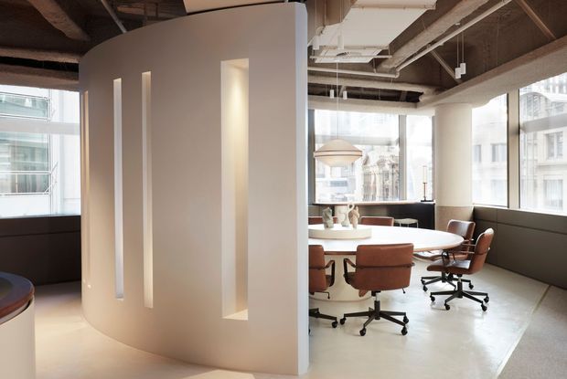 A bright contemporary commercial office interior with an open meeting area, a large round white conference table, rolling upholstered office chairs, expansive windows, and a sculptural curved partition wall featuring narrow recessed vertical openings. The exposed ceiling services, ductwork, and dark structural elements contrast with the clean light walls and polished pale floor.