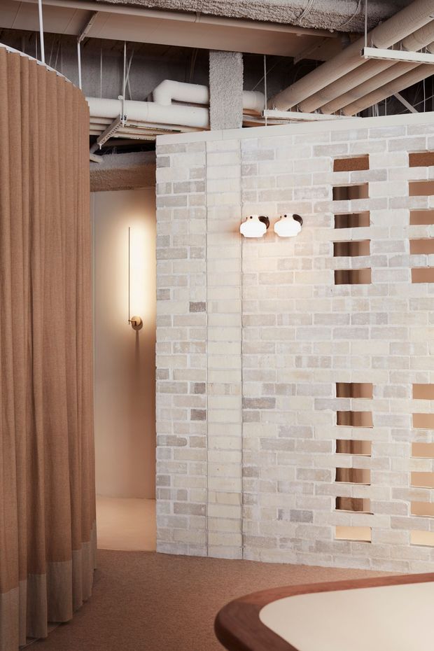 Interior view of a softly lit, industrial-style space featuring an exposed services ceiling with ducts and pipework, a pale brick breeze-block partition wall with horizontal openings, warm beige carpet, and a curved ribbed fabric curtain. A partially visible rounded timber-edged counter or table occupies the foreground.