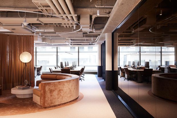 A contemporary commercial office reception and lounge area with a curved upholstered sofa, round side tables, decorative floor lamp, and a long communal worktable surrounded by office chairs. The space features exposed services and ductwork, a textured acoustic ceiling, full-height glazing, dark-framed glass partitions, warm timber slat screening, and a neutral beige and brown palette. A large mirror or reflective glass wall extends the visual depth of the office.