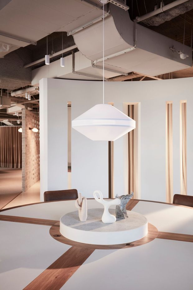 A contemporary commercial interior featuring a large communal dining or meeting table with a circular stone centre and timber radial inlay. A sculptural white pendant light hangs above the table. The space has pale partition walls with narrow recessed openings, exposed services and ductwork, brick columns, and a warm neutral palette with timber and leather accents.