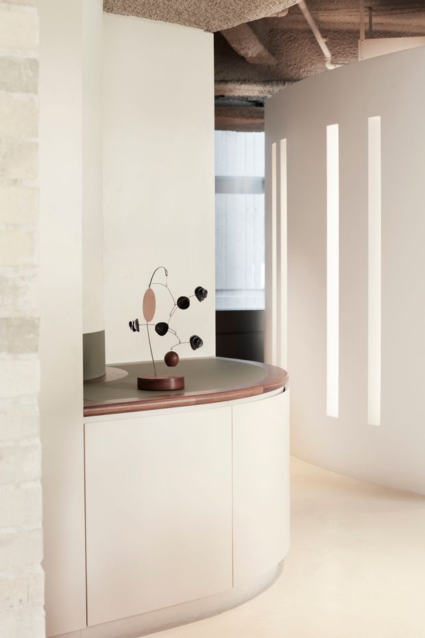 A softly lit minimalist interior vignette featuring a curved cream built-in cabinet with a muted green benchtop and reddish timber edge detail. A sculptural mid-century-style mobile sits on the counter, while narrow vertical openings in a curved partition wall cast bright bands of light. The space has exposed textured acoustic ceiling treatment, visible services, and a translucent window in the background.