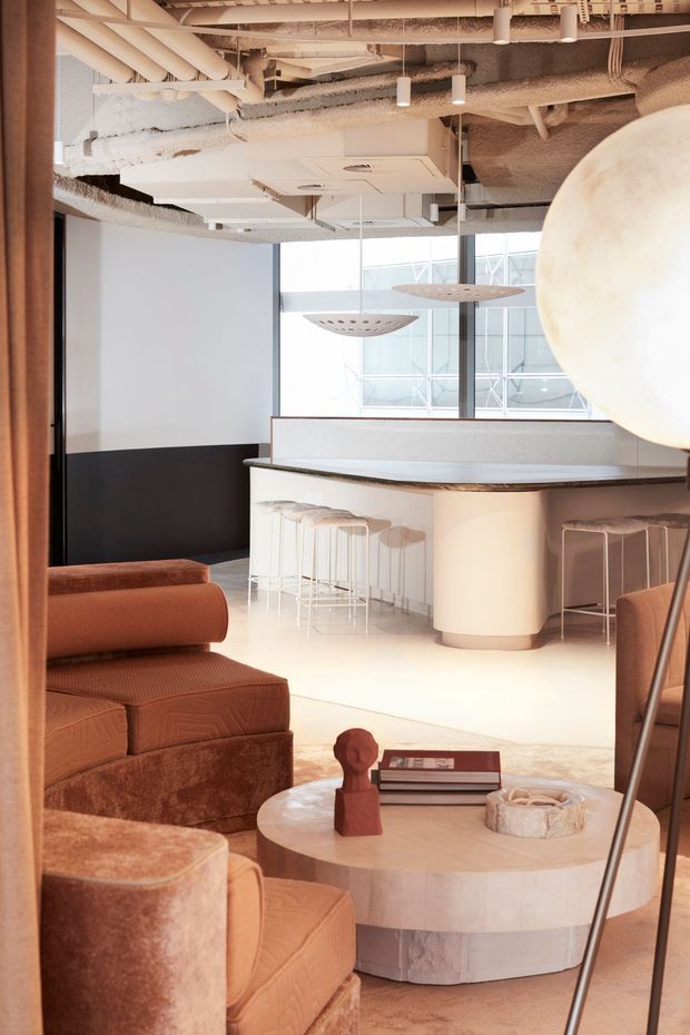 A stylish contemporary commercial lounge and bar interior with warm neutral tones. The foreground features plush rust-brown upholstered lounge chairs and a round pale stone coffee table with books and sculptural decor. Behind it is a curved white bar with a dark stone or timber-look benchtop, integrated seating, and transparent bar stools. Floor-to-ceiling glazing, sculptural pendant lights, and an exposed services ceiling create a refined hospitality or office-lounge atmosphere.