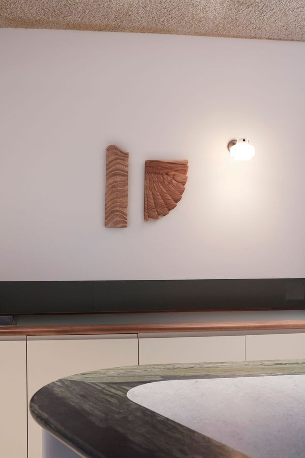 Close-up interior view of a refined built-in kitchen or bar area. A dark linear backsplash or cabinetry band runs across a pale wall above light cabinetry, with a warm timber ledge and a prominent curved stone benchtop in the foreground. Sculptural timber wall art and a small wall light add decorative detail. The ceiling has a textured finish.