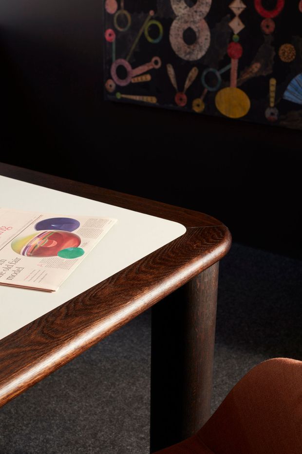 A close-up interior view of a dark, richly finished wooden table with rounded edges and a light inset tabletop surface. A magazine rests on the table, with a partial warm brown upholstered chair visible in the foreground. The room has dark walls, charcoal carpet, and a colourful abstract artwork mounted on the wall, creating a moody, refined atmosphere.