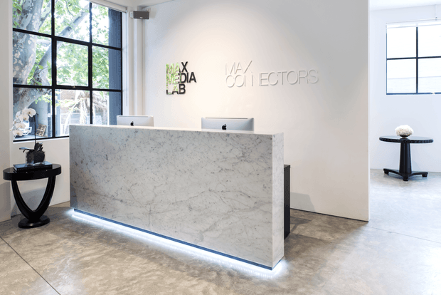 A bright modern commercial reception area featuring a large white veined stone reception desk with integrated underlighting, polished concrete flooring, black-framed floor-to-ceiling windows, and minimalist wall signage. A secondary open room with matching windows is visible beyond the reception space.