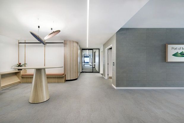 A bright, contemporary commercial reception or waiting area with a neutral grey carpet, white ceiling, and textured grey feature wall. The space includes a sculptural light-wood standing table, curved built-in timber bench seating, vertical timber slat detailing, pendant lights, and glazed doors leading to a corridor. A partial wall sign suggests an office or institutional setting.