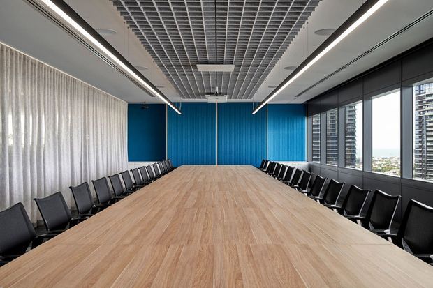 A large contemporary conference room with a long light-wood boardroom table surrounded by black chairs. The room features vivid blue ribbed feature walls, full-height sheer curtains, extensive windows overlooking nearby high-rise buildings, linear suspended lighting, and an exposed white grid ceiling.