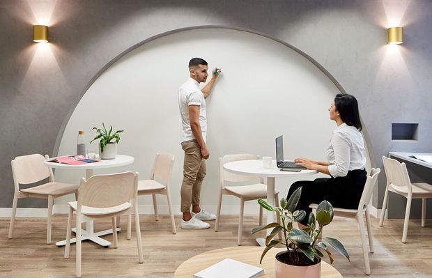 A contemporary collaborative workspace or café-style office with pale timber tables and chairs, a textured grey wall, warm wall sconces, and a large arched feature opening. Two people are working around tables, with a laptop, whiteboard activity, plants, and a clean light-toned interior.