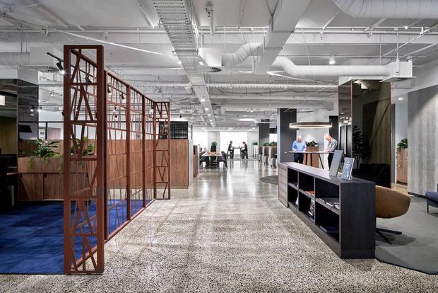 A bright, open-plan commercial interior with an industrial-style exposed services ceiling, polished terrazzo-like flooring, timber and glass meeting screens, built-in reception or display joinery, and collaborative work areas. The space is predominantly white, grey, black, and warm timber, with a refined contemporary workplace fit-out.