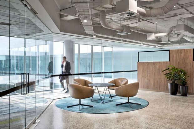 A contemporary commercial office reception or breakout lounge with floor-to-ceiling glass partitions, exposed services and ductwork, polished concrete flooring, a wood-panel feature wall, and a circular seating arrangement around a small table on a blue rug. The space is bright and open, with views through extensive glazing and indoor plants.