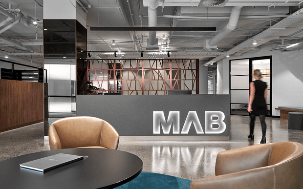 A contemporary commercial reception and waiting area with a large illuminated MAB reception sign, black reception desk, exposed services ceiling, polished concrete floor, timber and black metal partitions, and tan leather lounge chairs around a round black table.