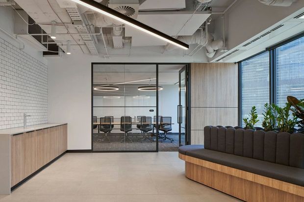 Contemporary commercial office reception or waiting area with a built-in curved timber reception bench upholstered in dark brown, pale concrete-look flooring, white brickwork, timber wall panelling, black-framed glazed meeting-room partitions, and abundant indoor planting. The ceiling has exposed services, ducts, suspended linear lighting, and circular pendant fixtures, with a meeting room visible beyond.